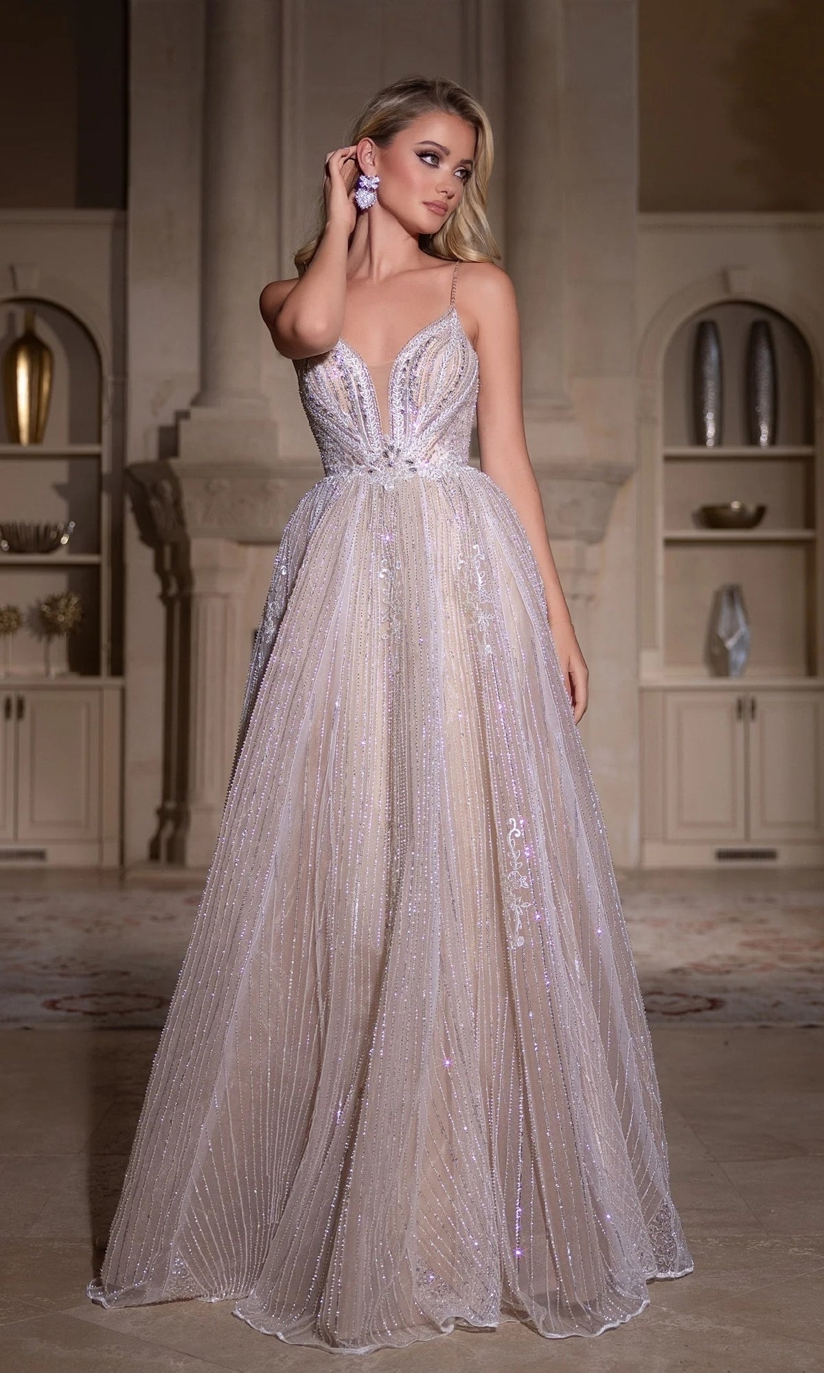 A woman stands indoors, touching her earring, wearing the Portia and Scarlett PS25310 long prom dress—a floor-length, light-colored gown with beaded embellishments, a plunging neckline, and sparkling details.